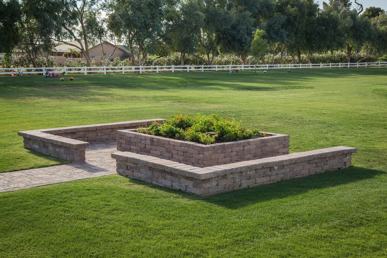 A tiered stone planter box with greenery sits on a grassy lawn near a paved path, framed by a white fence and trees.