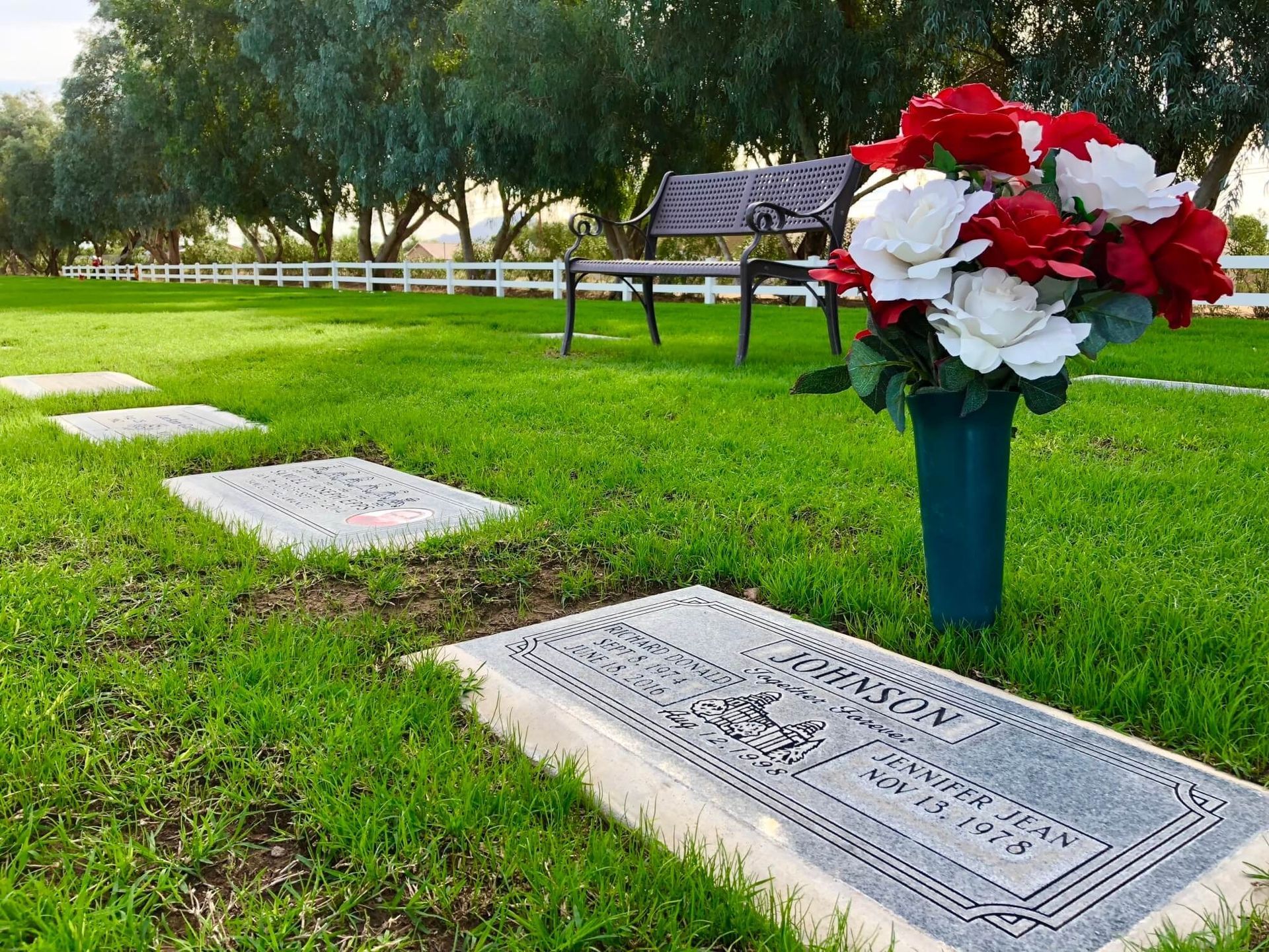 A cemetery lawn with flat headstones, a bench in the background, and a bouquet of red and white flowers in a green vase.