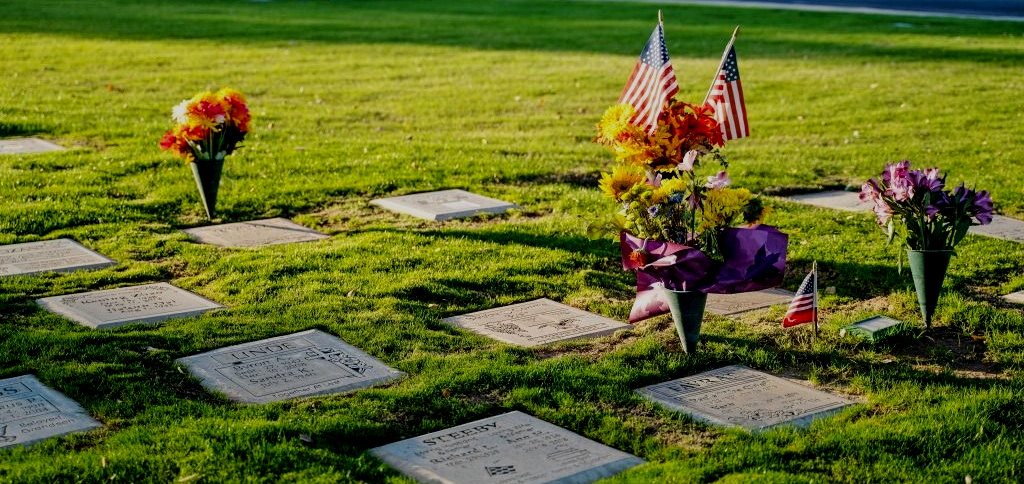 Sunlit cemetery with rows of flat grave markers, bouquets of flowers, and small American flags at sunset.