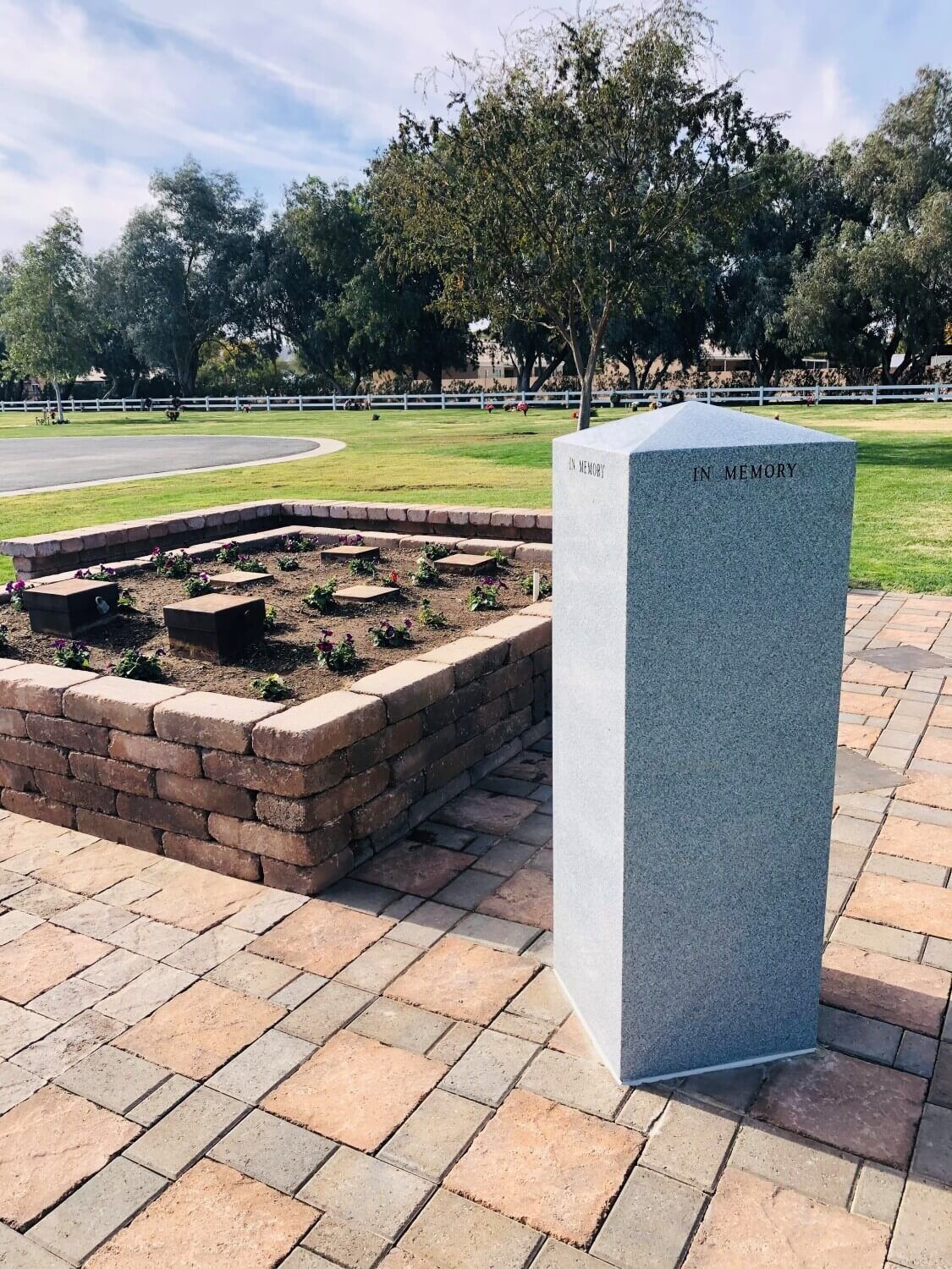 A granite monument stands on a paved patio next to a brick-walled garden bed in a grassy park setting.
