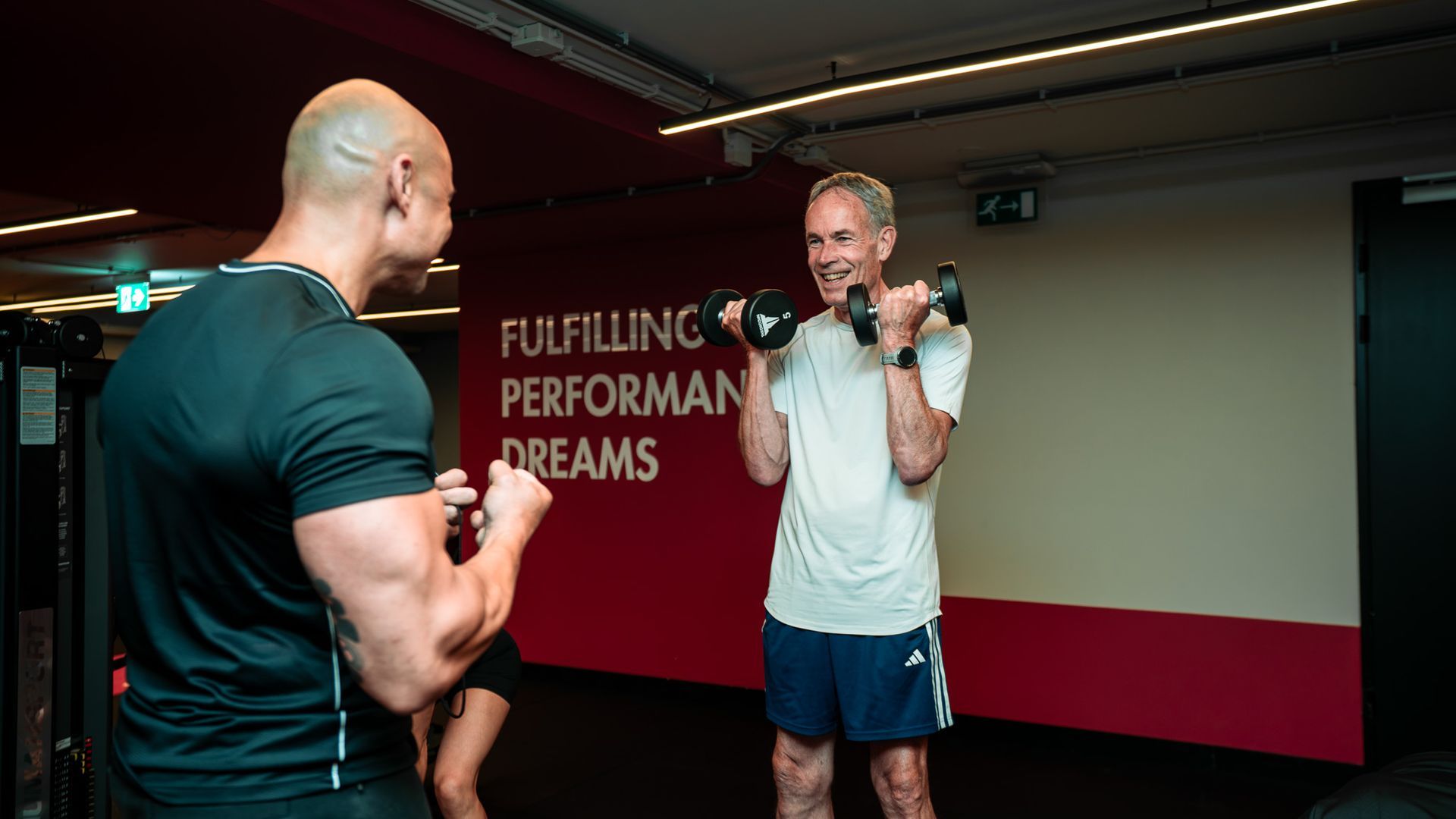 Een trainer begeleidt een zittende cliënt in een sportschool, met fitnessapparatuur op de achtergrond.