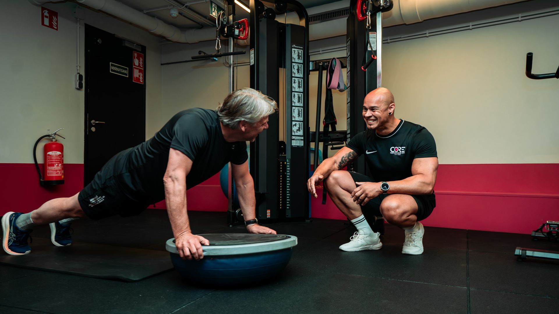 Een man hangt aan de ringen in een schemerige, industriële fitnessruimte, gezien door metalen tralies.