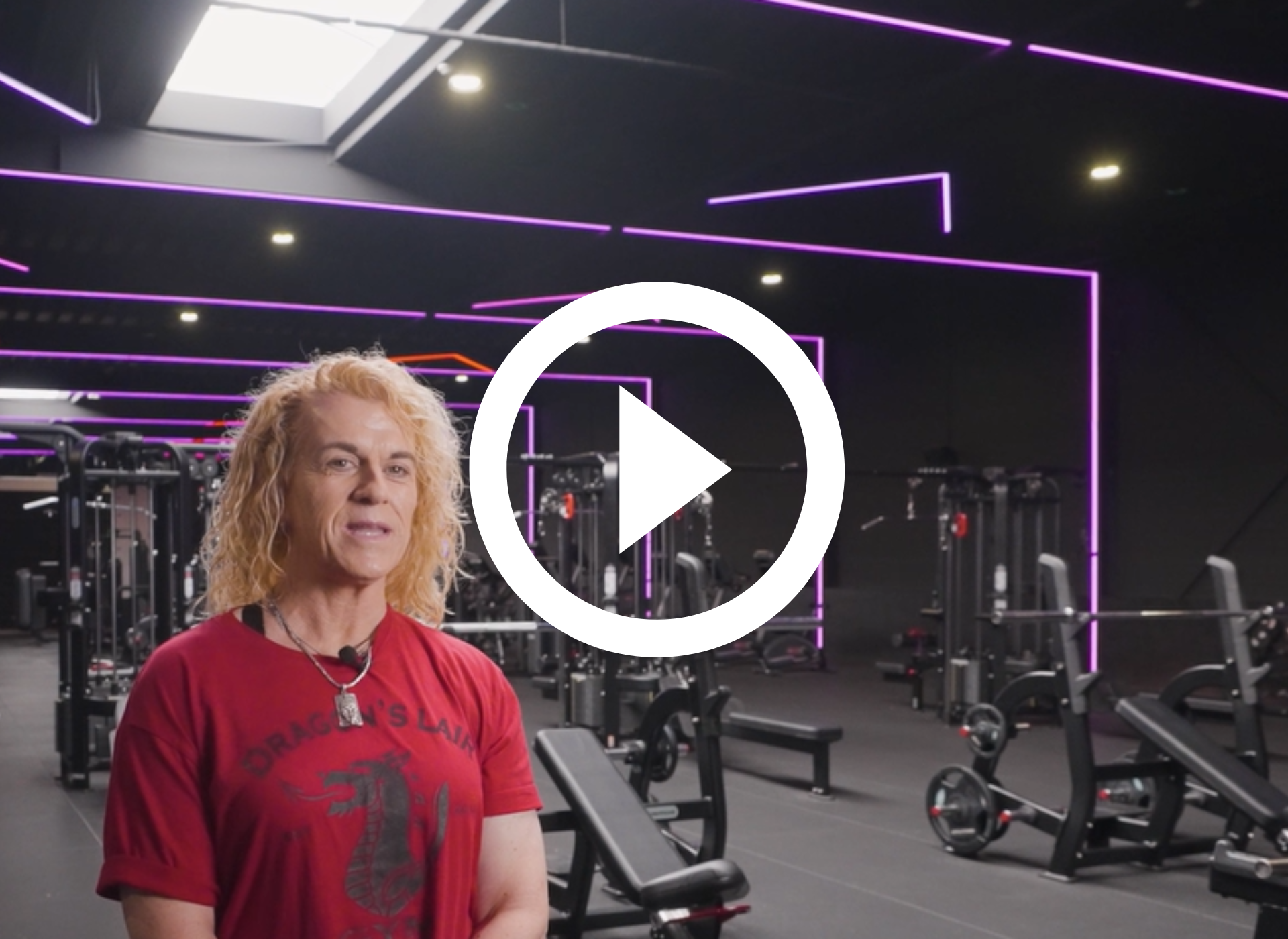 Een man in een rood shirt staat in een sportschool met neonverlichting, fitnessapparaten en een grote afspeelknop.