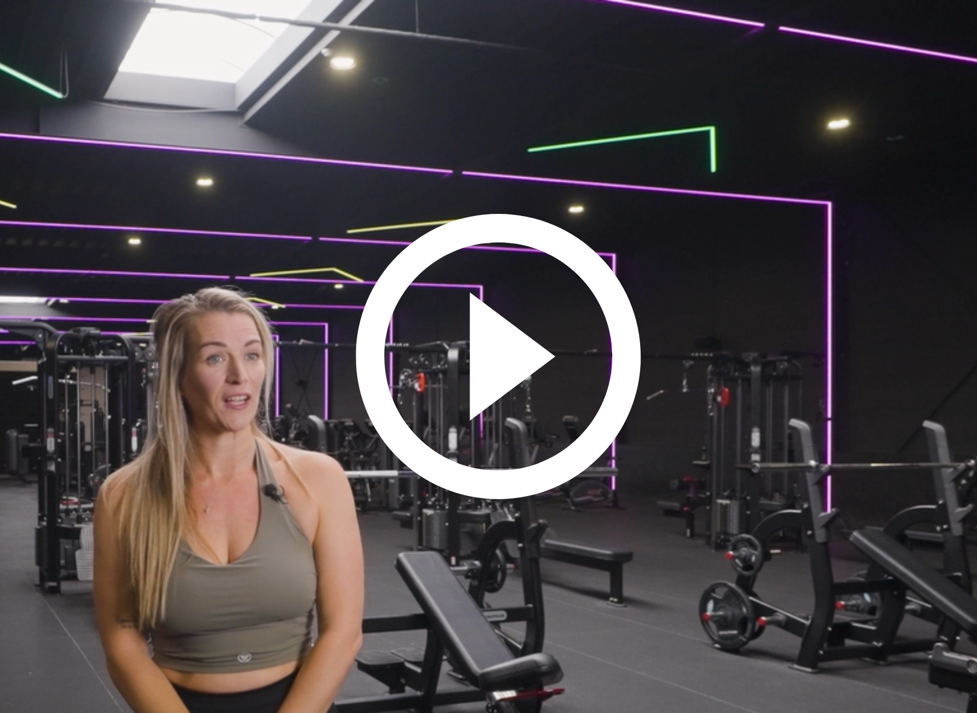 Vrouw in een sportschool met fitnessapparaten en neonverlichting aan het plafond, met daarop een afspeelknop.