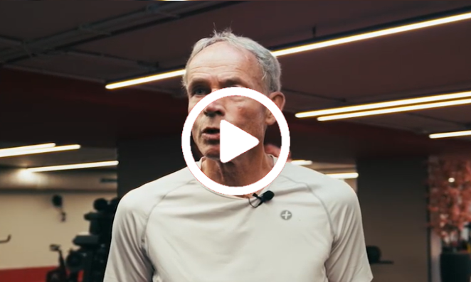 Een man in een rood shirt staat in een sportschool met neonverlichting, fitnessapparaten en een grote afspeelknop.