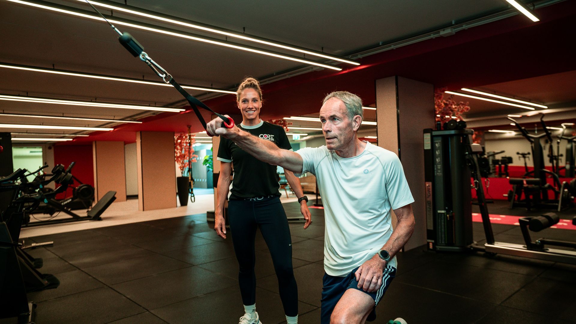 Een trainer helpt een vrouw met sit-ups in een sportschool, met dumbbells op de achtergrond.