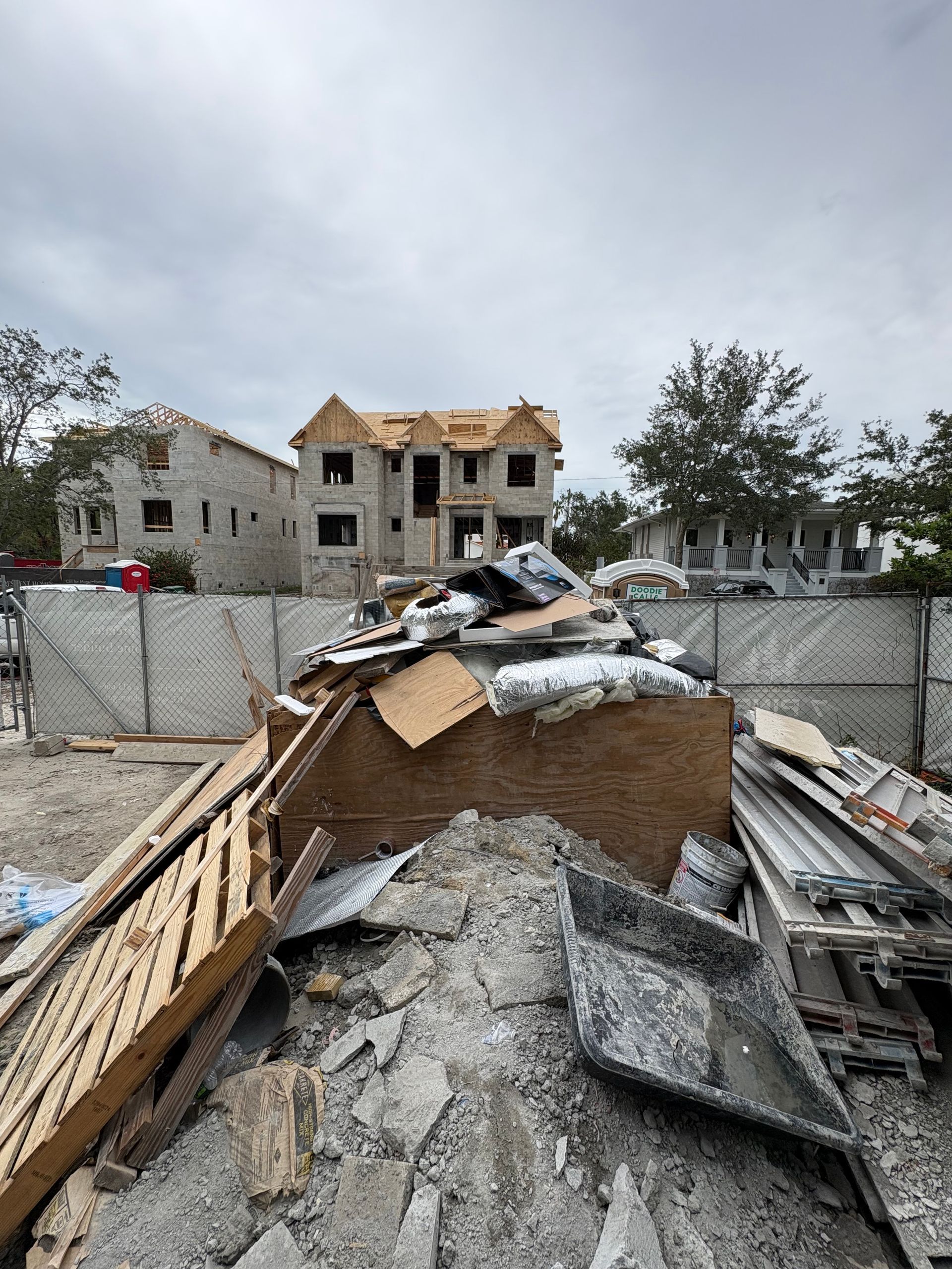 Collapsed debris in a fenced lot with damaged apartment buildings in the background under a gray sky
