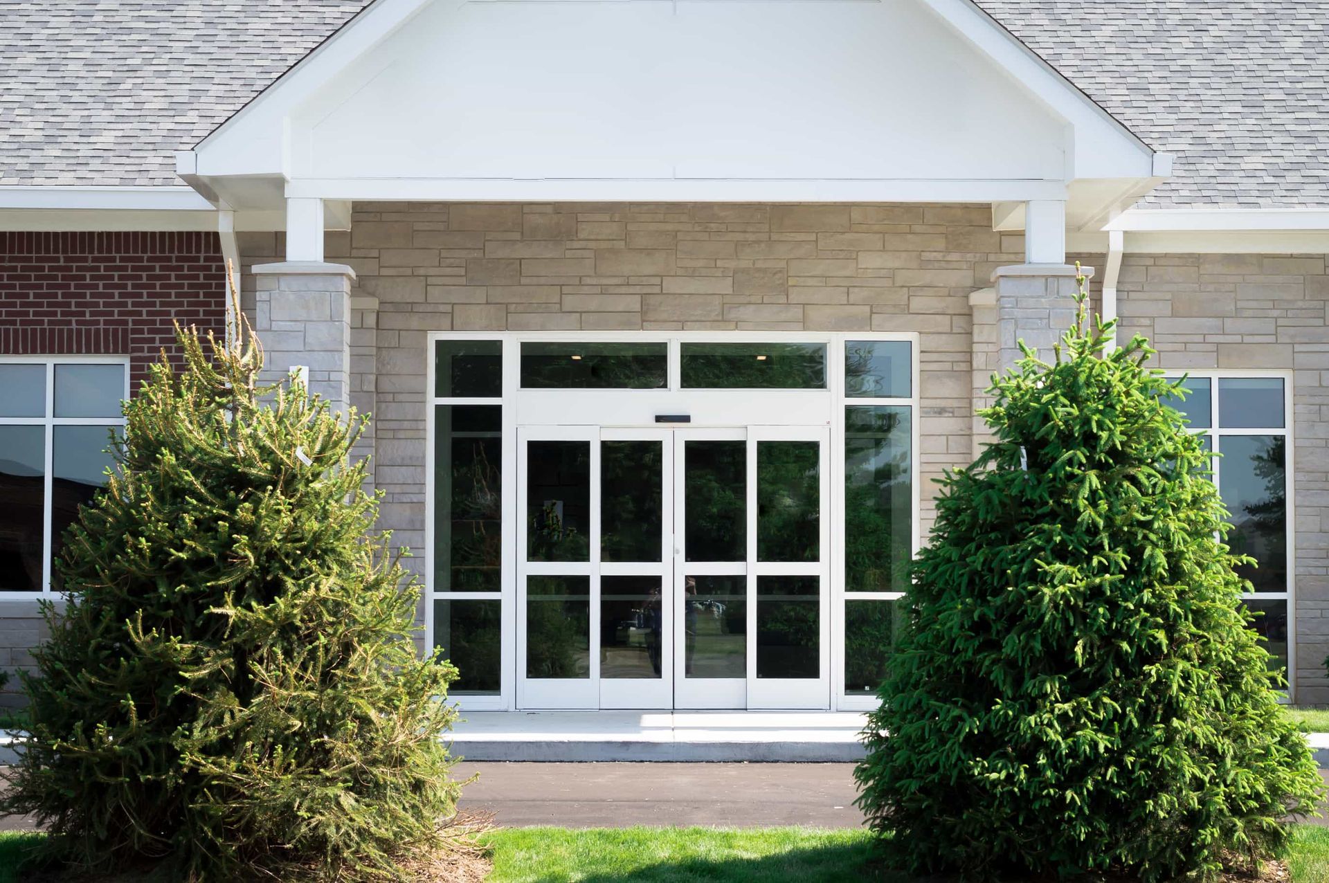 Front entrance of a building with glass doors, white columns, and two large evergreen shrubs.