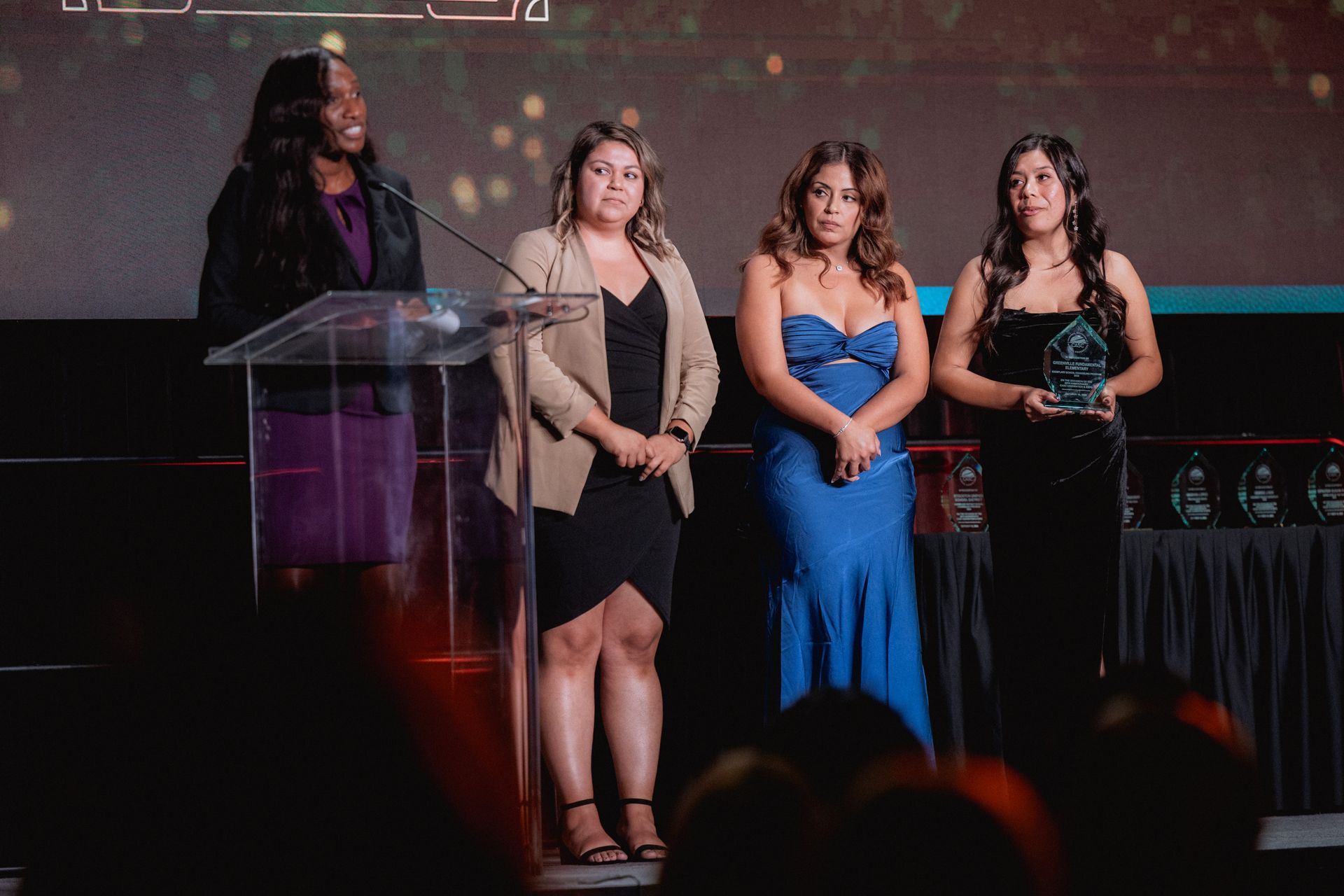 Four people on stage at an awards ceremony. One speaks at a podium; others stand, holding an award.