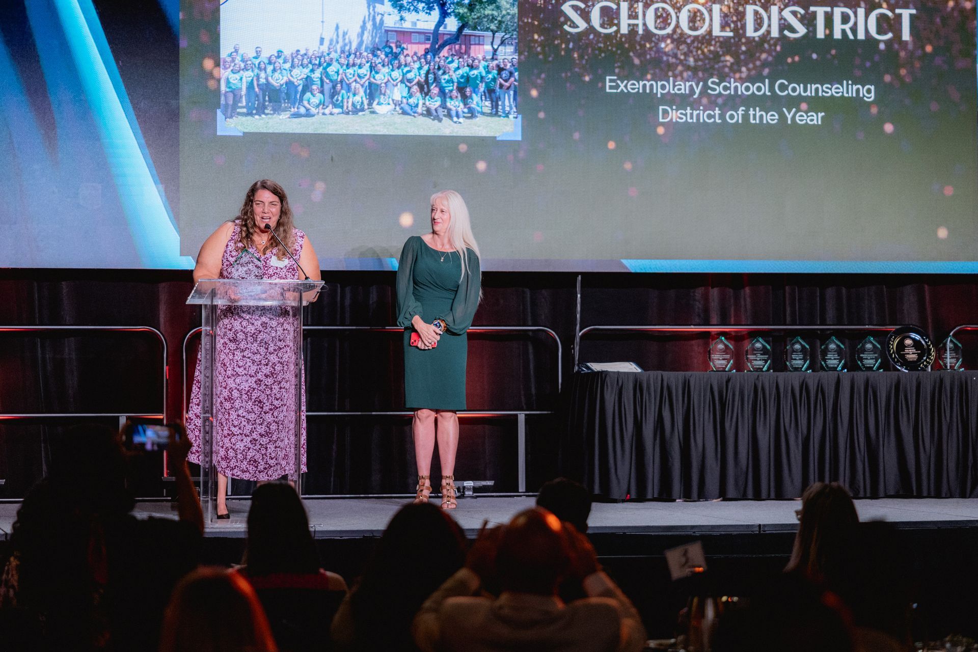 Two women on stage at an awards ceremony, one speaking at a podium, the other standing.