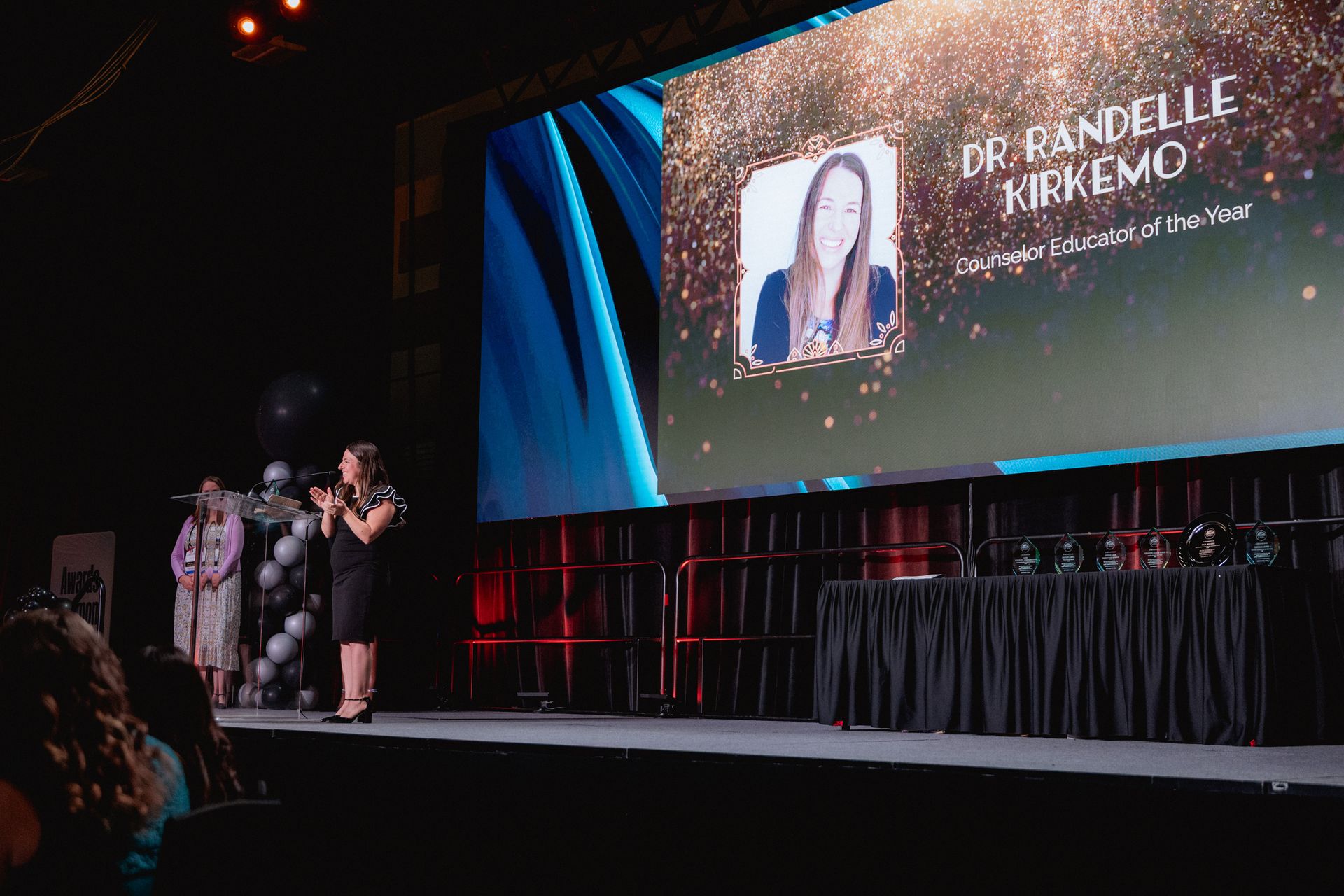 Woman on stage at awards event, screen displays 