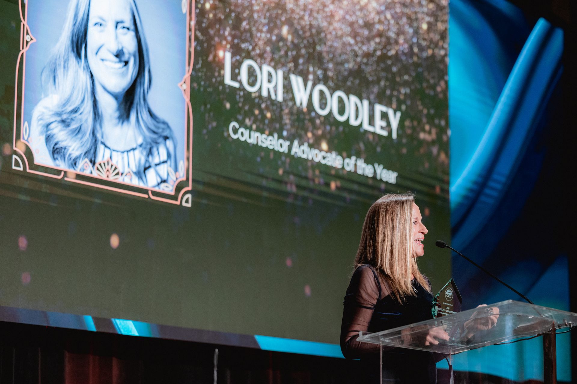Lori Woodley, named Counselor Advocate of the Year, speaks at podium with screen of her photo.