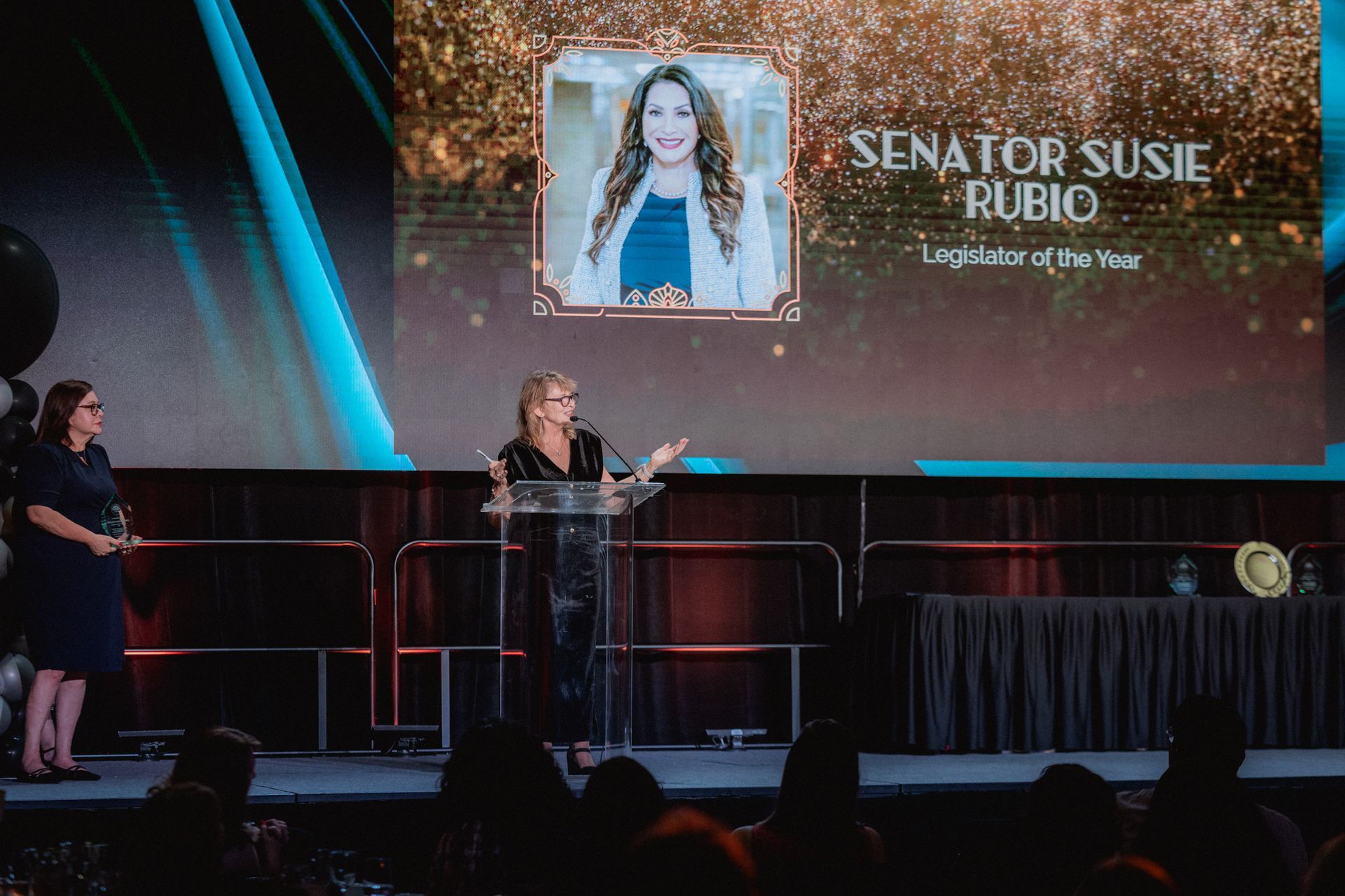 Woman speaking at podium, with a screen behind her displaying 