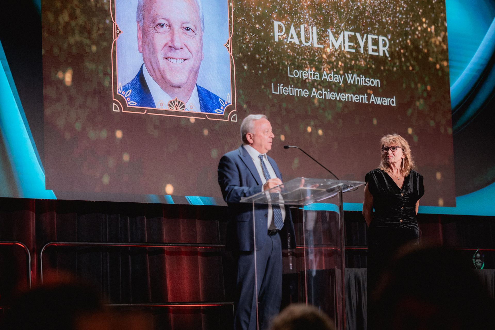 Man at podium with woman beside him, accepting an award. A screen behind them shows the man's photo and name.