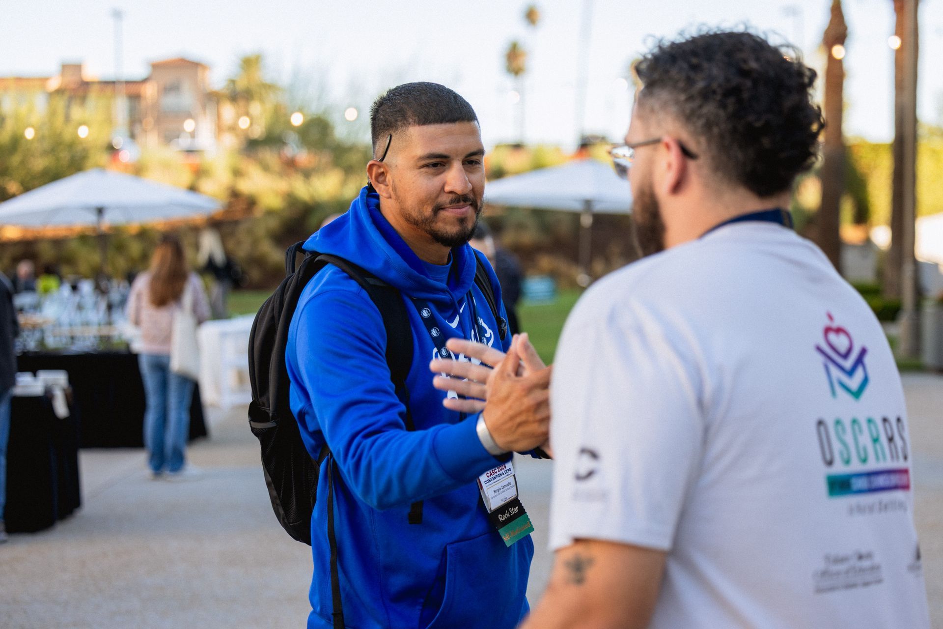 Two men shake hands outdoors. One wears a blue hoodie and backpack. The other wears a white shirt with a logo.