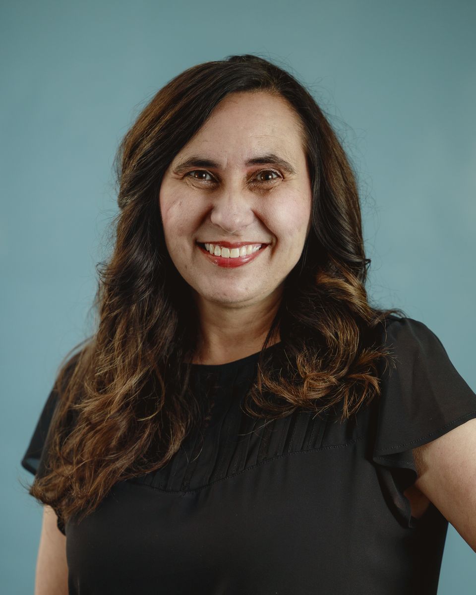 Smiling person with long dark hair in a black shirt against a teal background