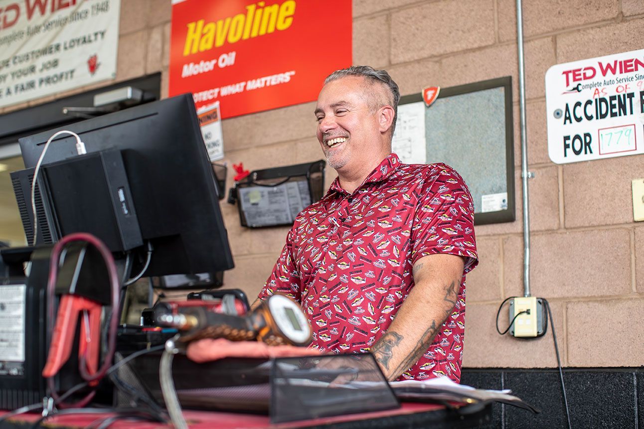 Man smiling at a desk in a garage, using a computer near a Havoline sign.