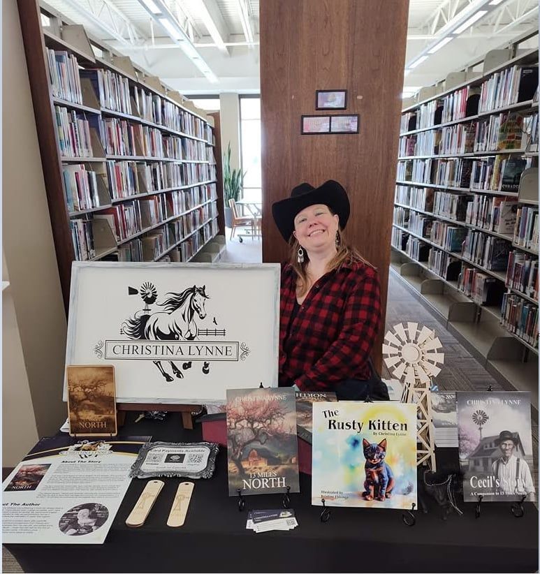 An author wearing a cowboy hat smiles behind a table displaying her books, a wooden windmill, and signs in a library.
