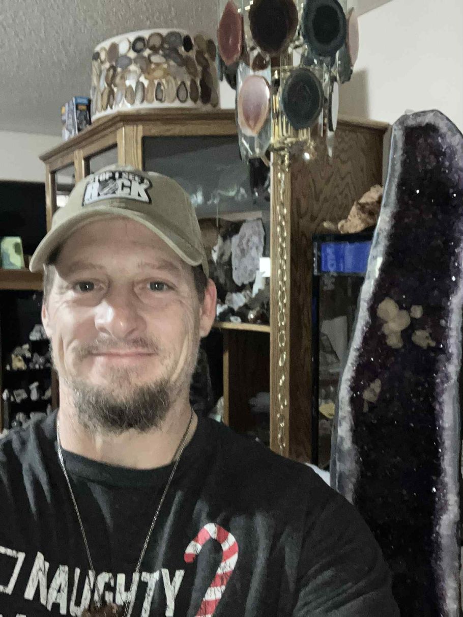 A person with a beard and baseball cap posing in front of a tall, dark amethyst crystal formation in a home interior.