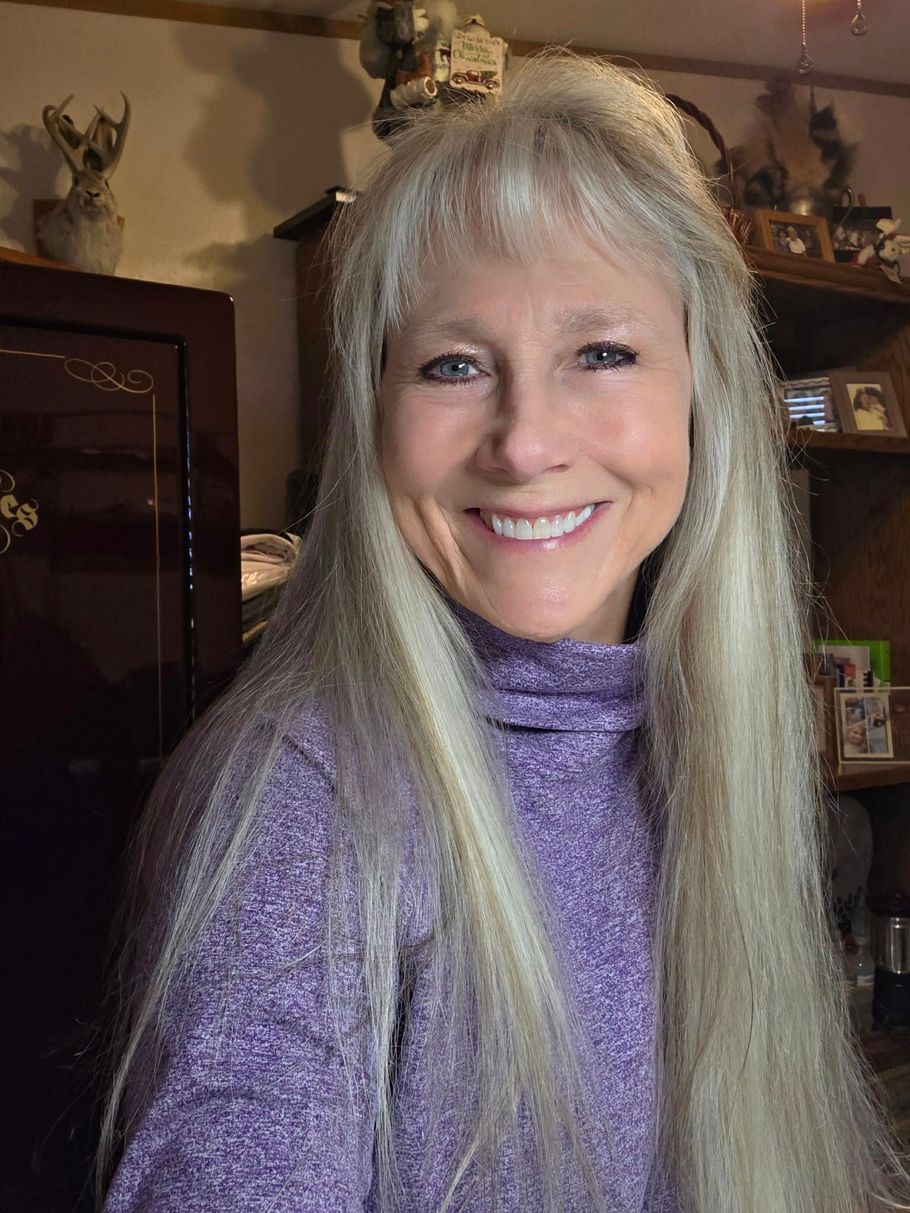 A smiling person with long, wavy blonde hair and bangs, wearing a purple mock-neck sweater indoors.