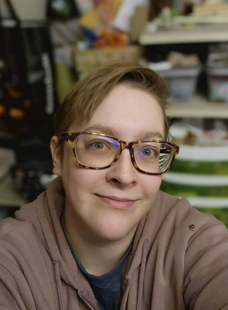 A person with short brown hair wearing tortoise-shell glasses and a dusty-pink hoodie, smiling at the camera.