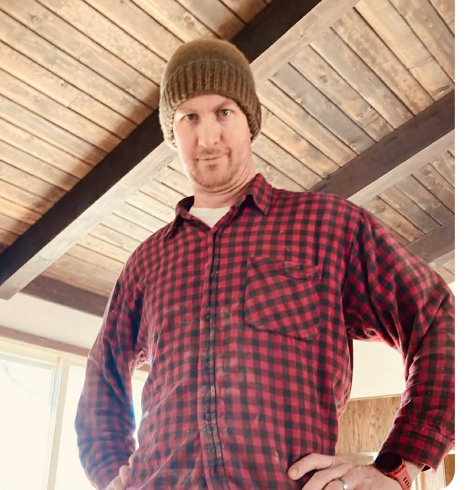A person wearing a brown beanie and red plaid flannel shirt stands with hands on hips under a wooden ceiling.