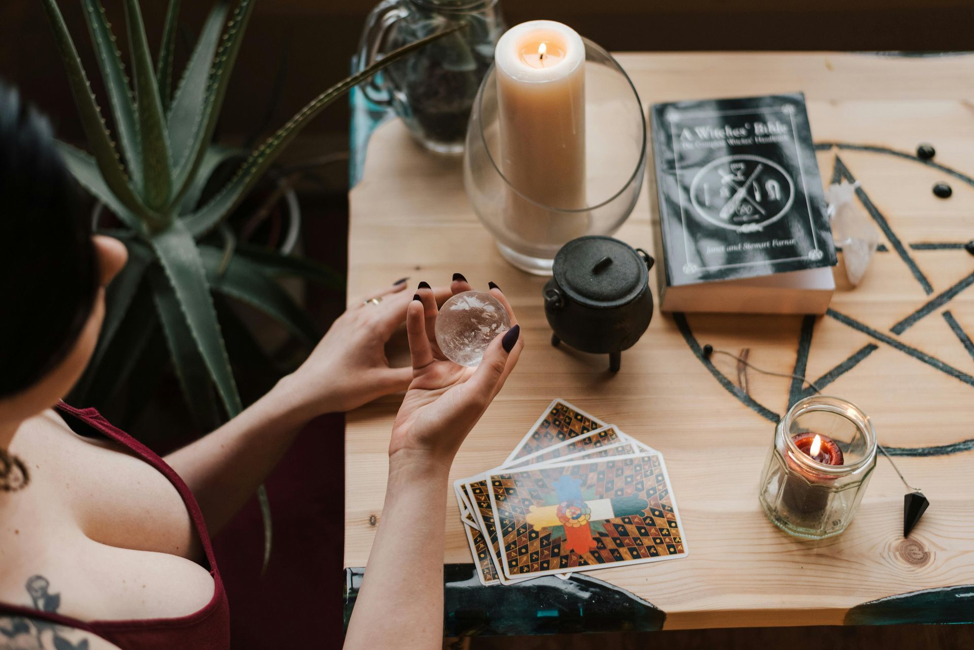 Woman holding crystal ball, cards, candles, book, and plant on wooden table.