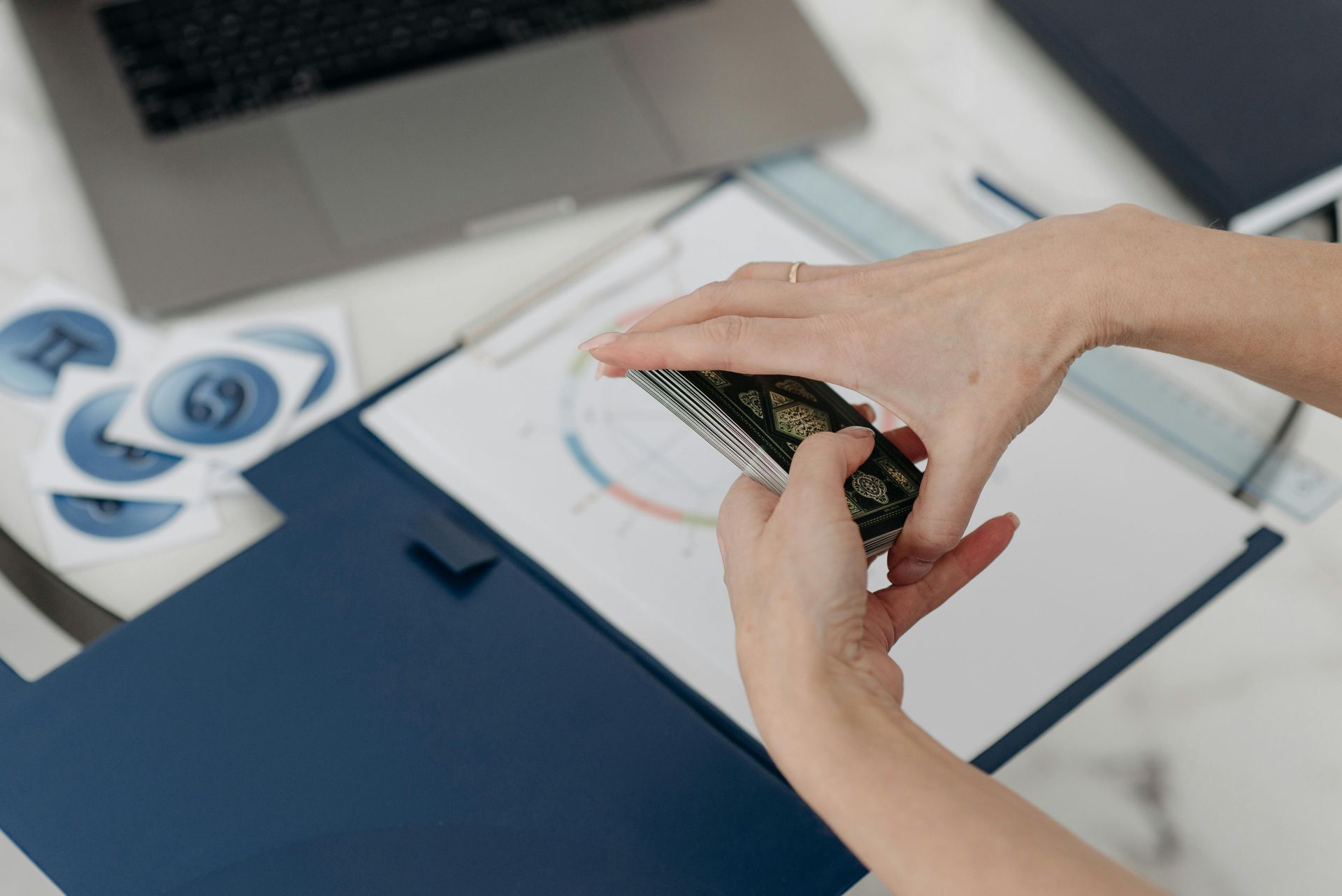 Hands shuffle a deck of tarot cards over an astrological chart on a desk with a laptop and zodiac stickers.
