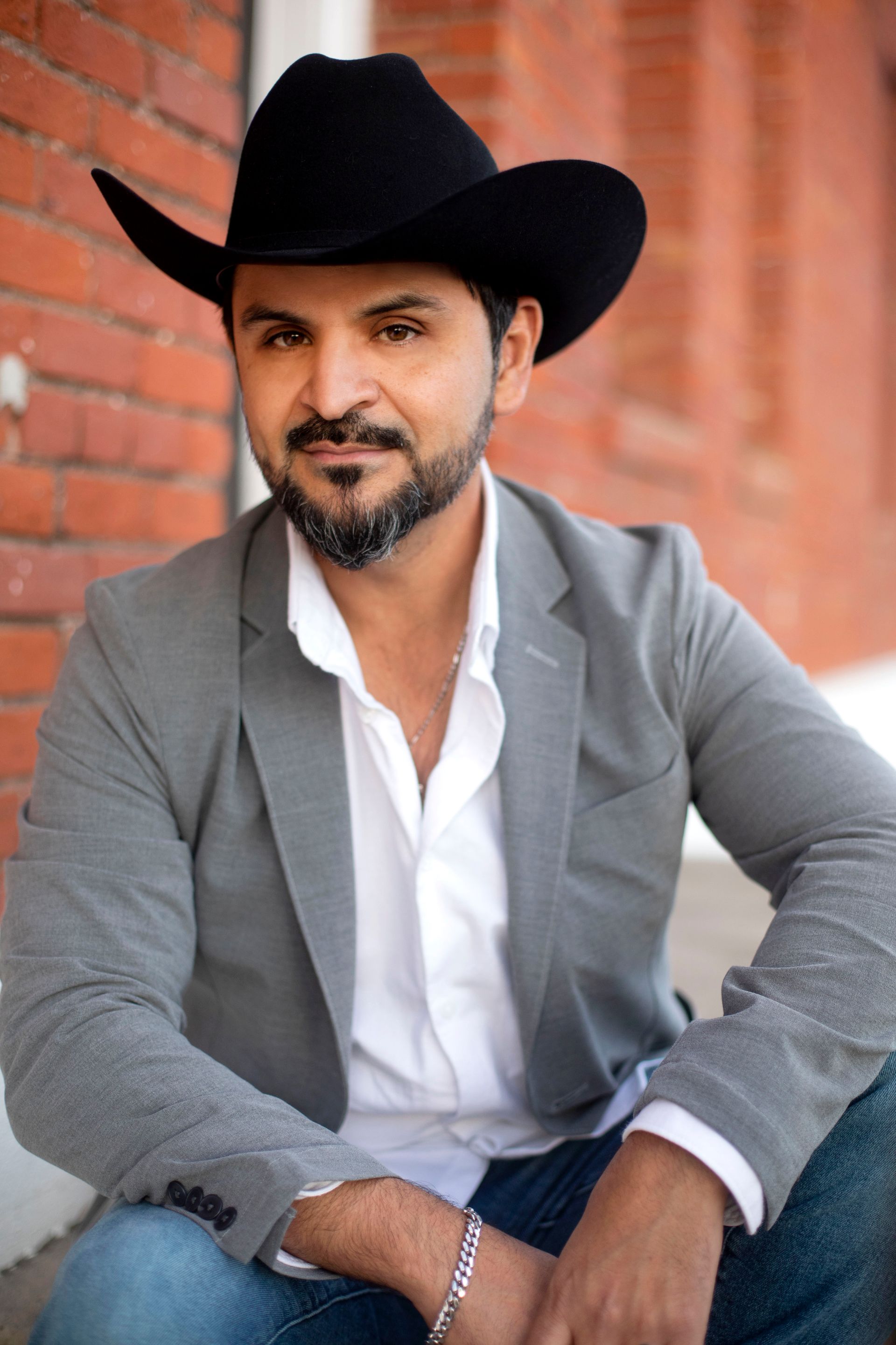 A man wearing a cowboy hat and a gray jacket is sitting in front of a brick wall.