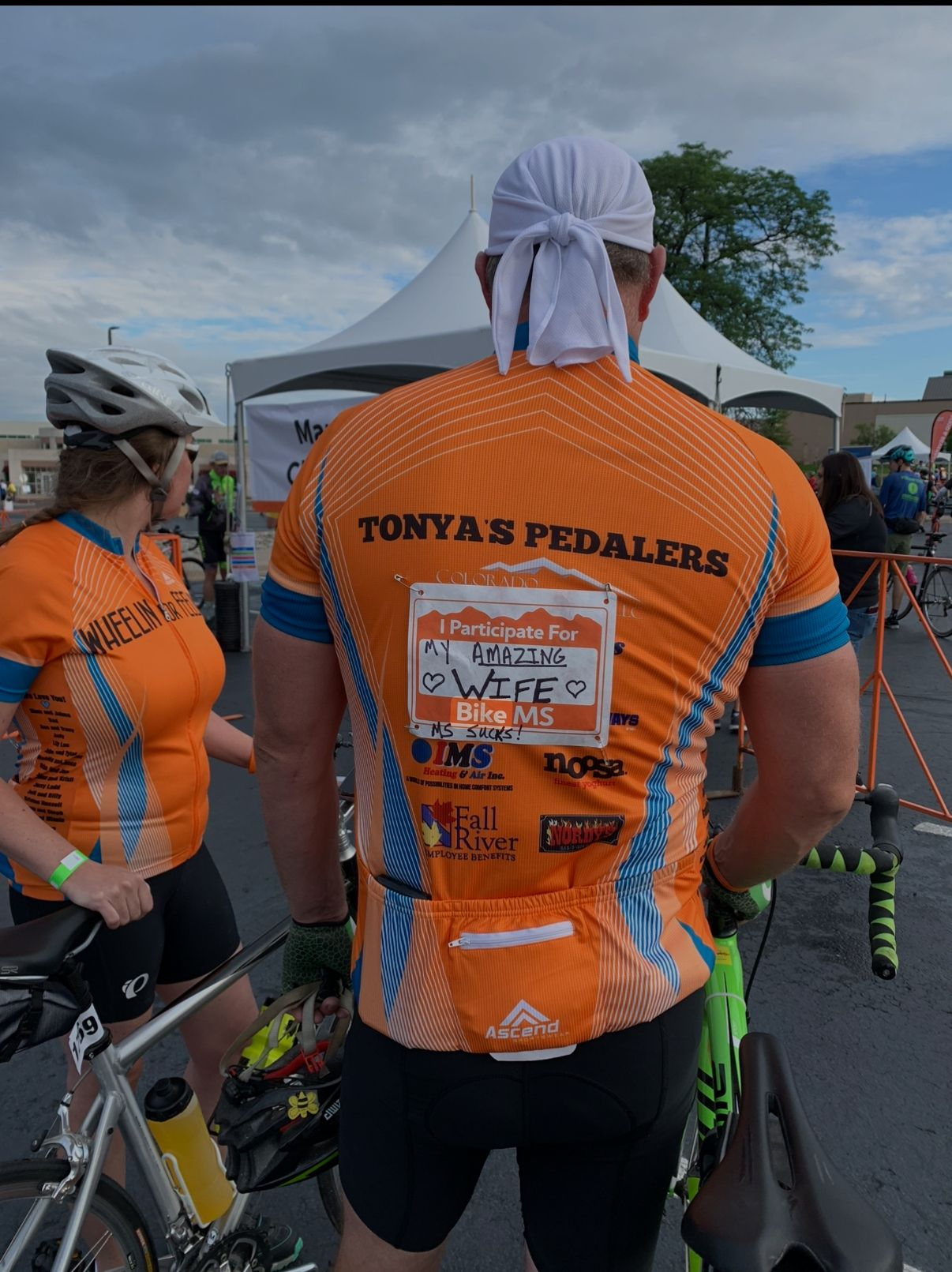 Two cyclists in orange jerseys at an outdoor event. One wears a white head covering.