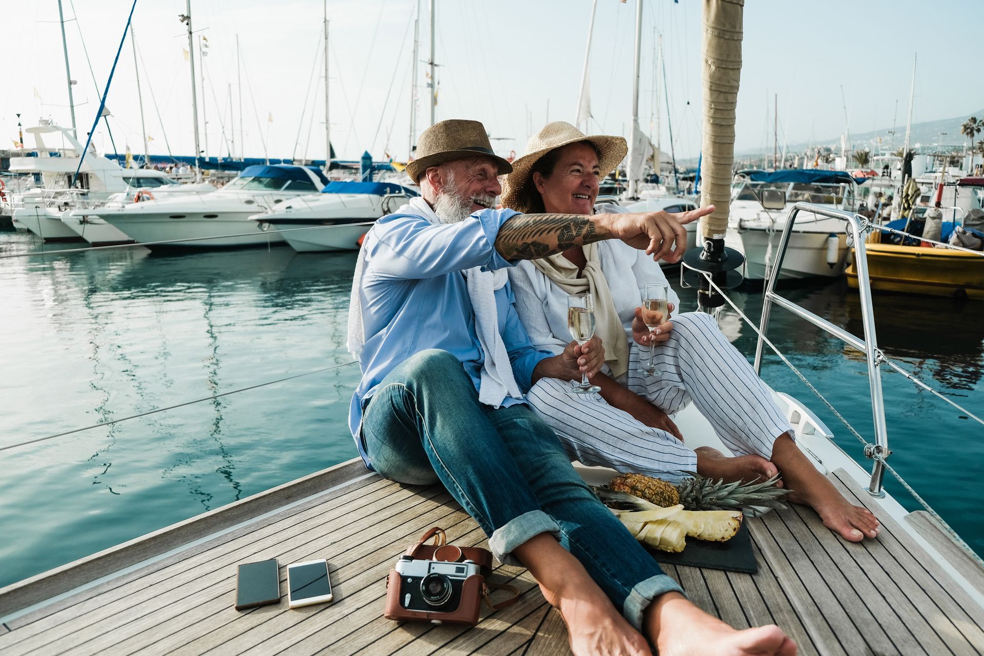 Couple on yacht, man points, woman smiles. Boats in background. Water and sky.