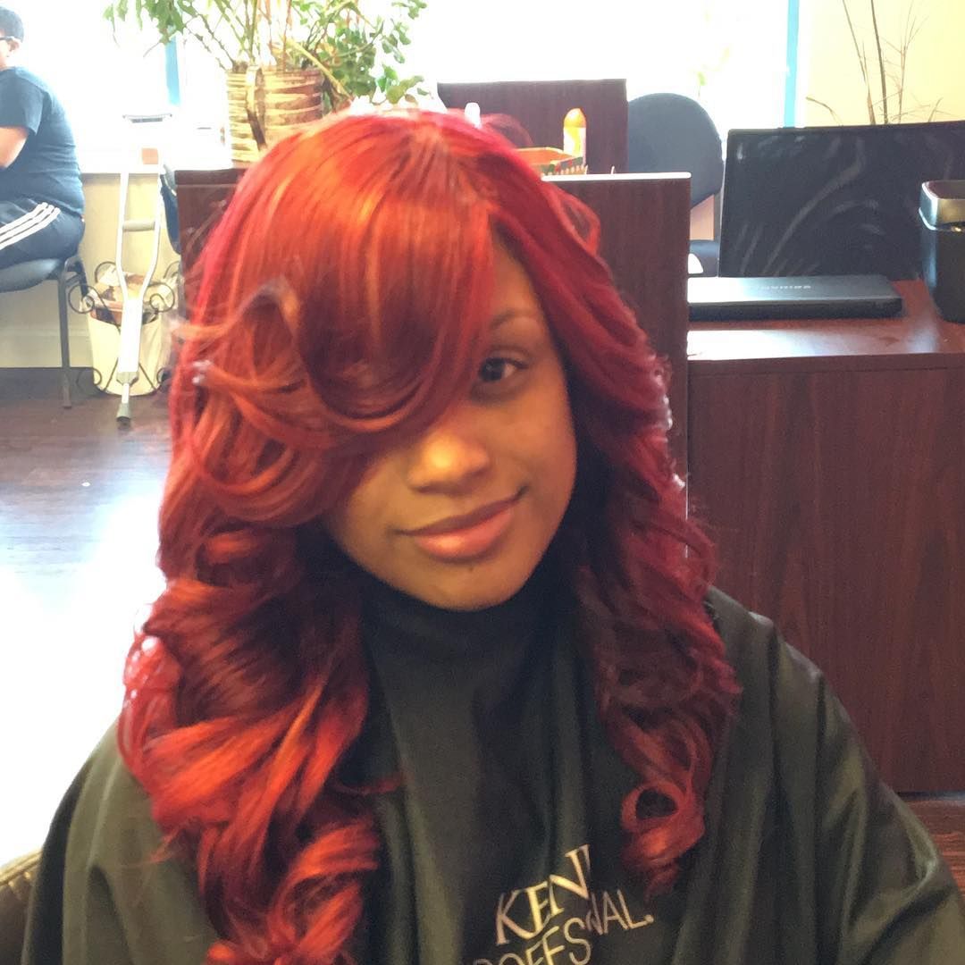 Woman with bright red curls and side-swept bangs, smiling indoors at a salon chair.