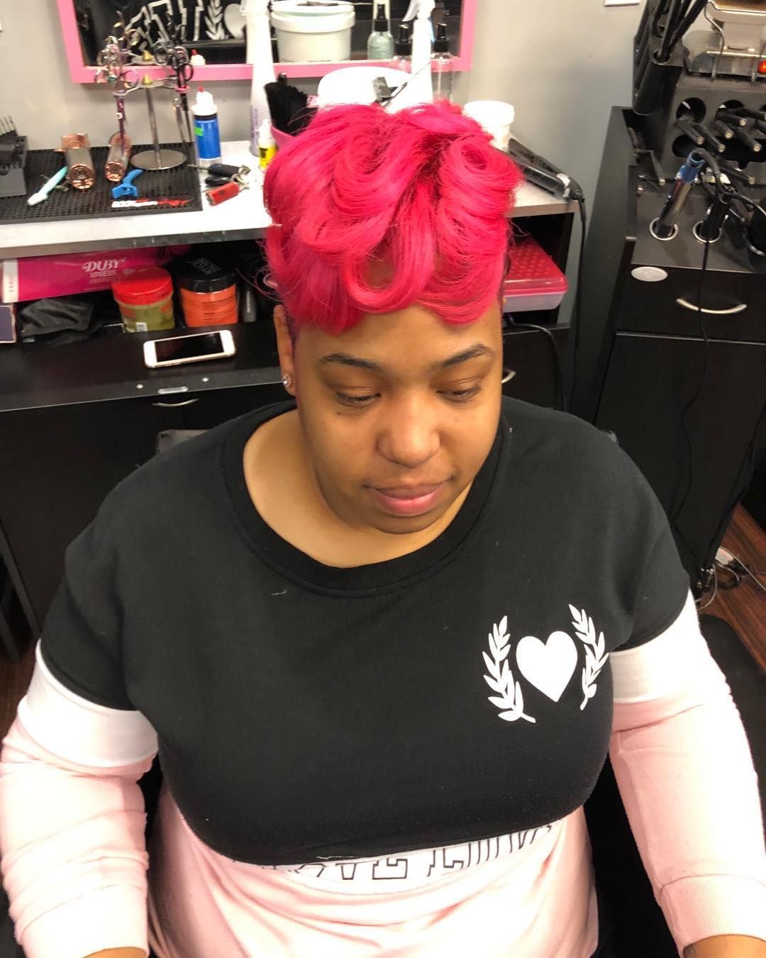 Person with bright pink hair sitting in a salon chair, looking down, wearing a black shirt over white sleeves.