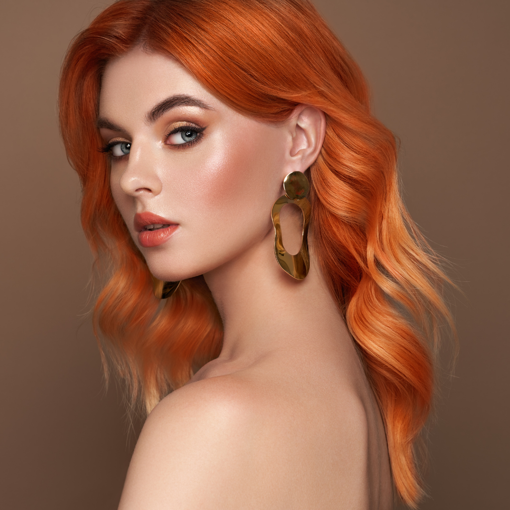 Portrait of a woman with wavy orange hair and gold earrings against a neutral background