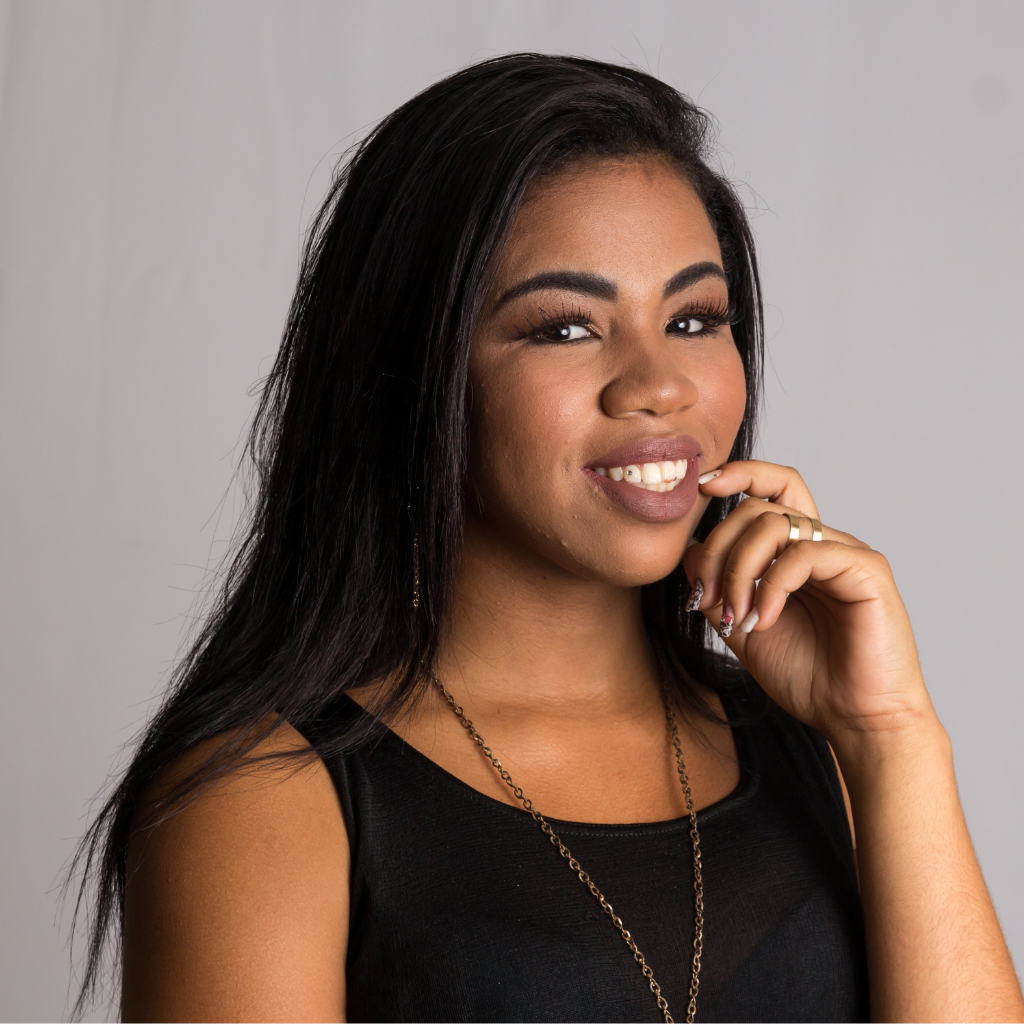Smiling woman in a black top, resting her hand on her chin against a plain gray background