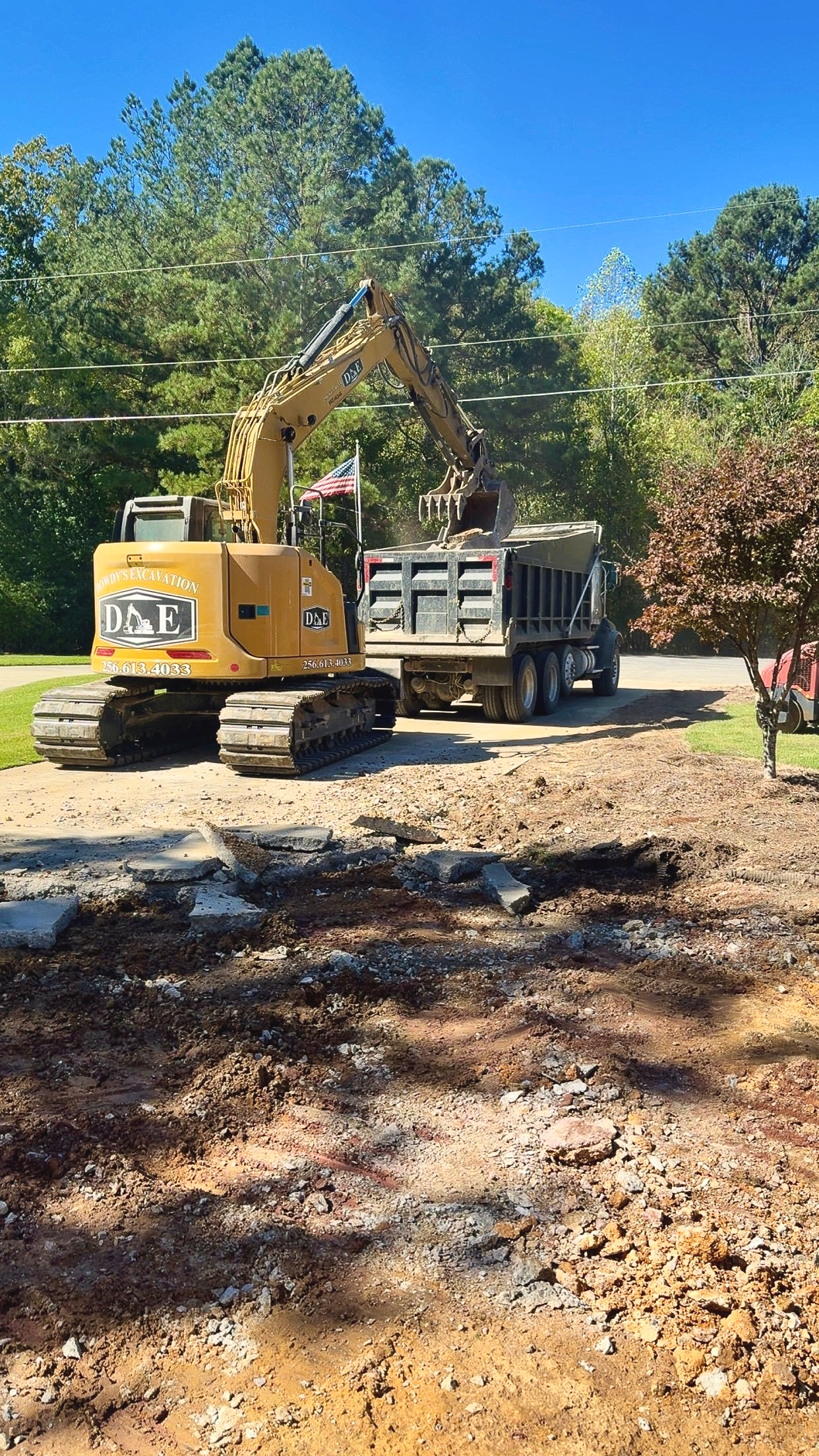 A yellow Caterpillar excavator loads dirt and debris into the back of a dump truck on a dirt road near trees.