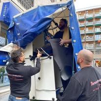 Tre operai sollevano un telone blu sopra un macchinario in una corsia di un negozio.