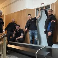 Quattro persone in posa in interni, accanto a una parete e una scala in legno; una di loro è accovacciata e fa il segno della pace.