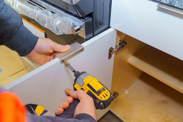 Mani che utilizzano un trapano giallo per installare una cerniera in un mobile da cucina grigio.