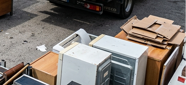 Elettrodomestici e cartone dismessi ammucchiati su un carrello accanto a un camion in strada.
