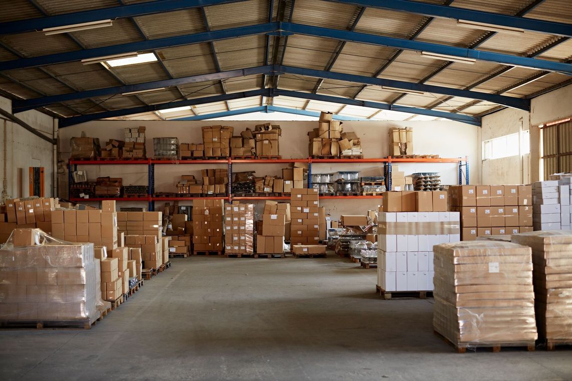 Interno di un magazzino con scatole di cartone e pallet impilati sotto un alto tetto metallico.