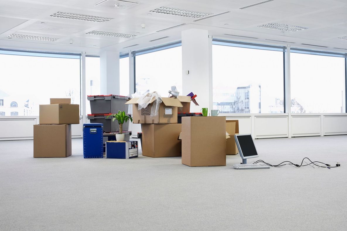 Scatole e materiali per il trasloco sparsi in un ufficio luminoso e vuoto con grandi finestre.