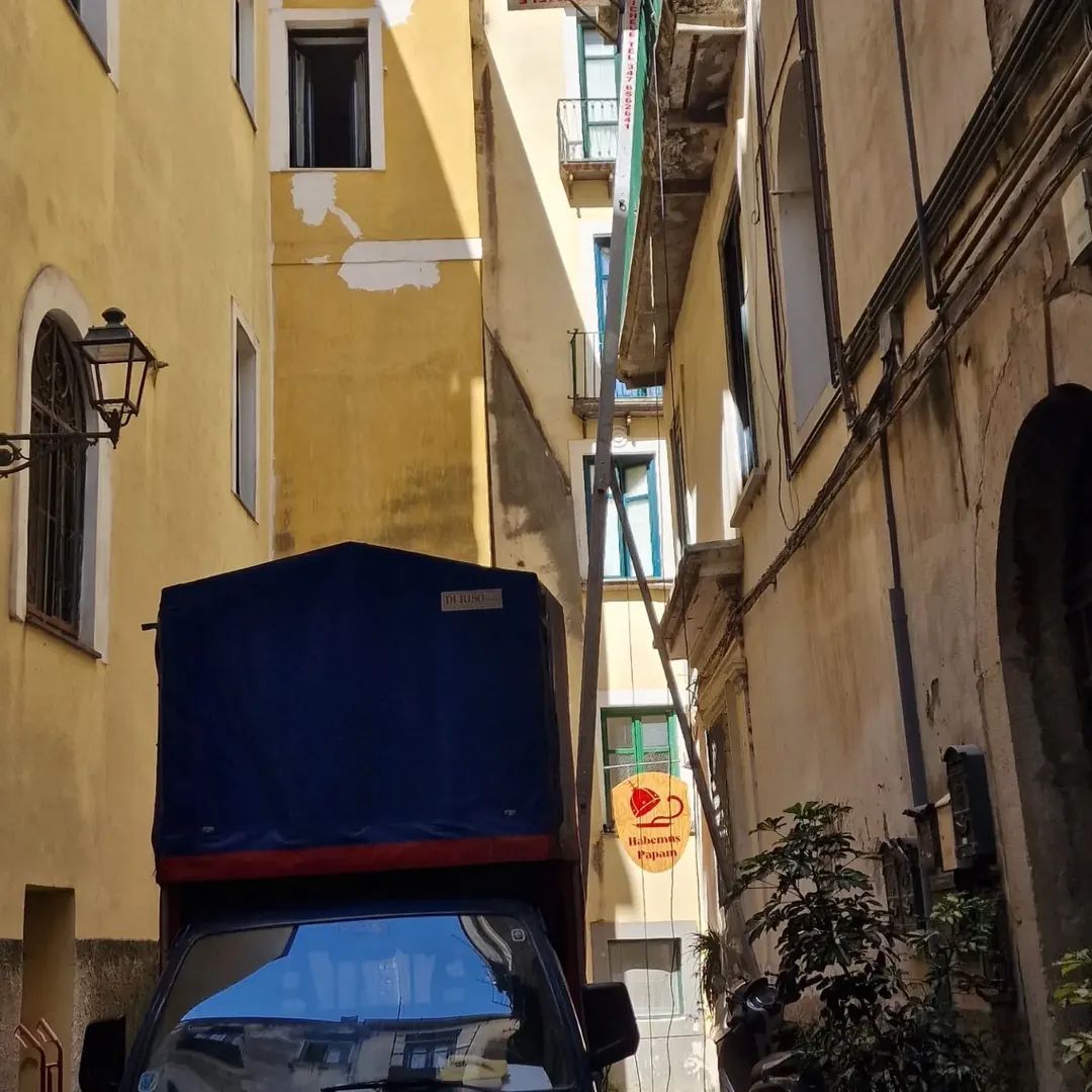 Stretto vicolo tra edifici gialli e beige, con un camion blu parcheggiato sotto un cartello appeso.