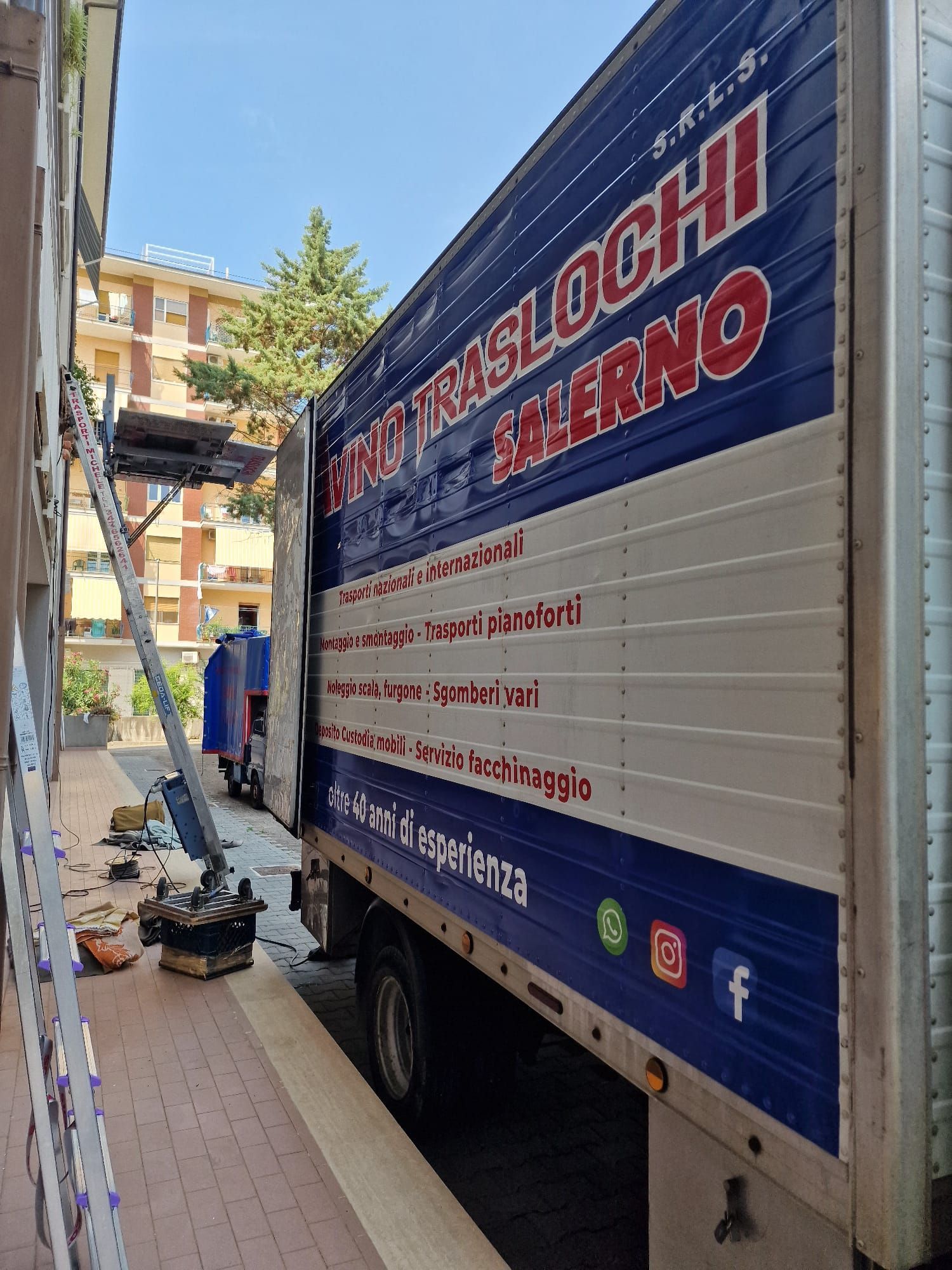 Un camion per traslochi è parcheggiato su uno stretto marciapiede accanto a un edificio, con attrezzi e scatole a terra.