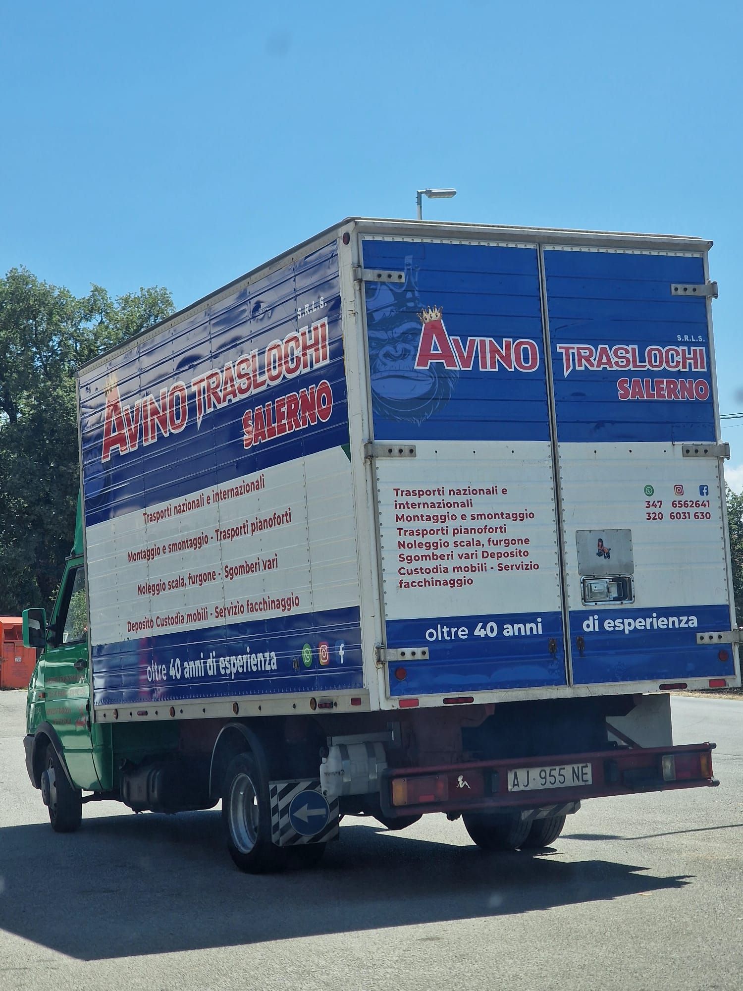 Un camion per traslochi blu e bianco con scritte rosse che percorre una strada soleggiata.