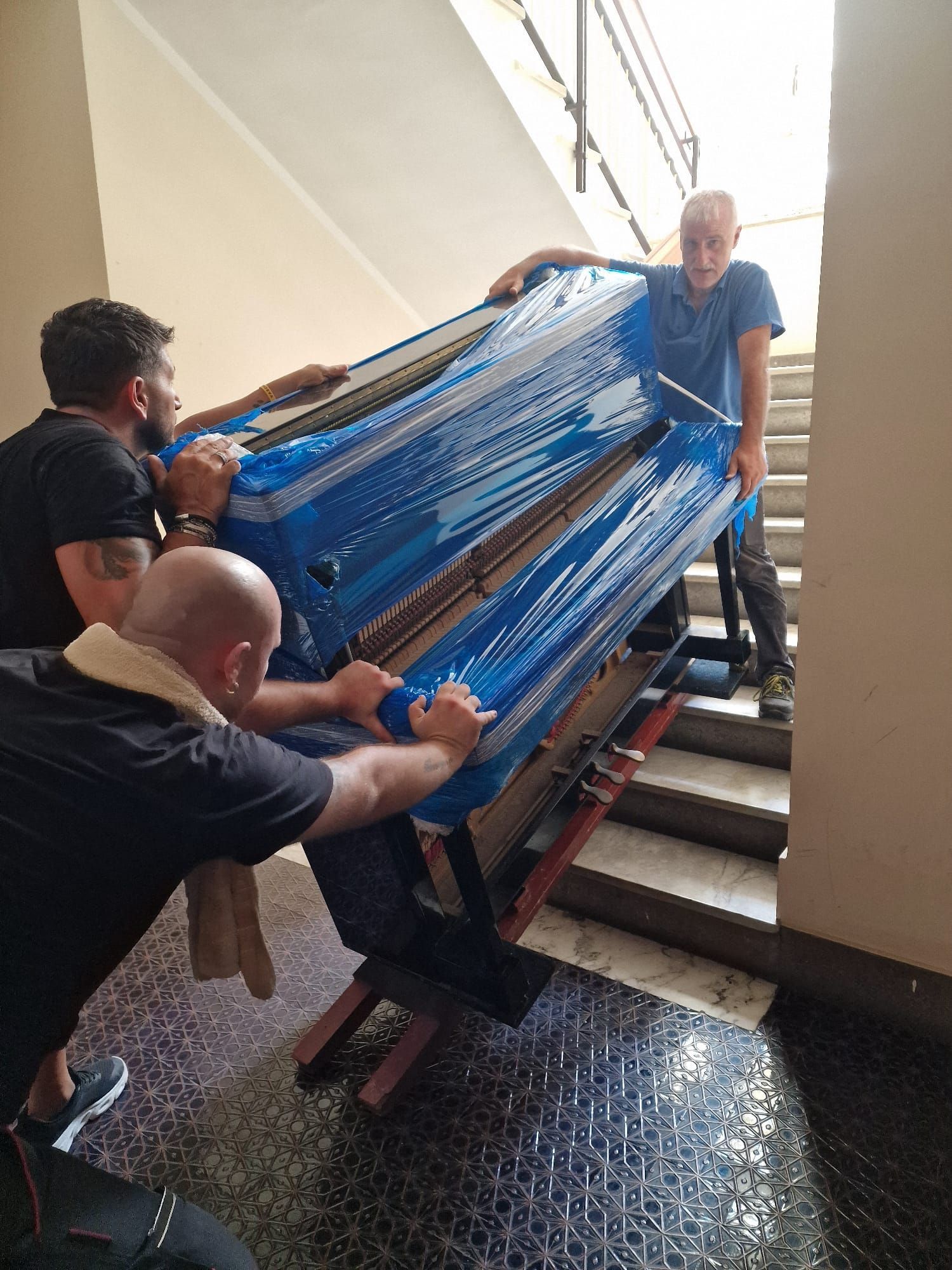 Tre operai trasportano un grande oggetto avvolto in un telo blu su per una stretta tromba delle scale.
