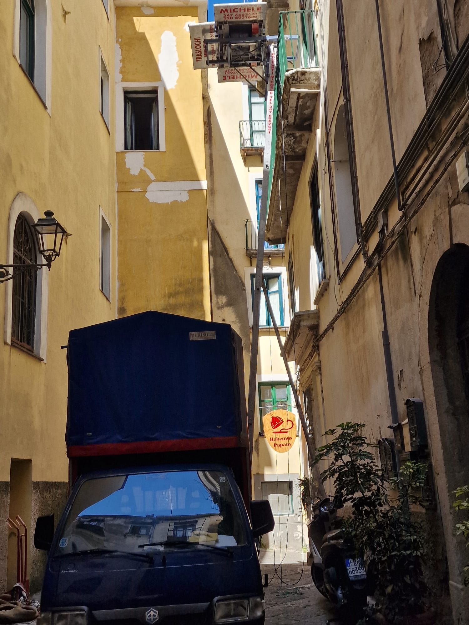 Stretto vicolo con edifici alti e un furgone parcheggiato che trasporta un cassone blu.