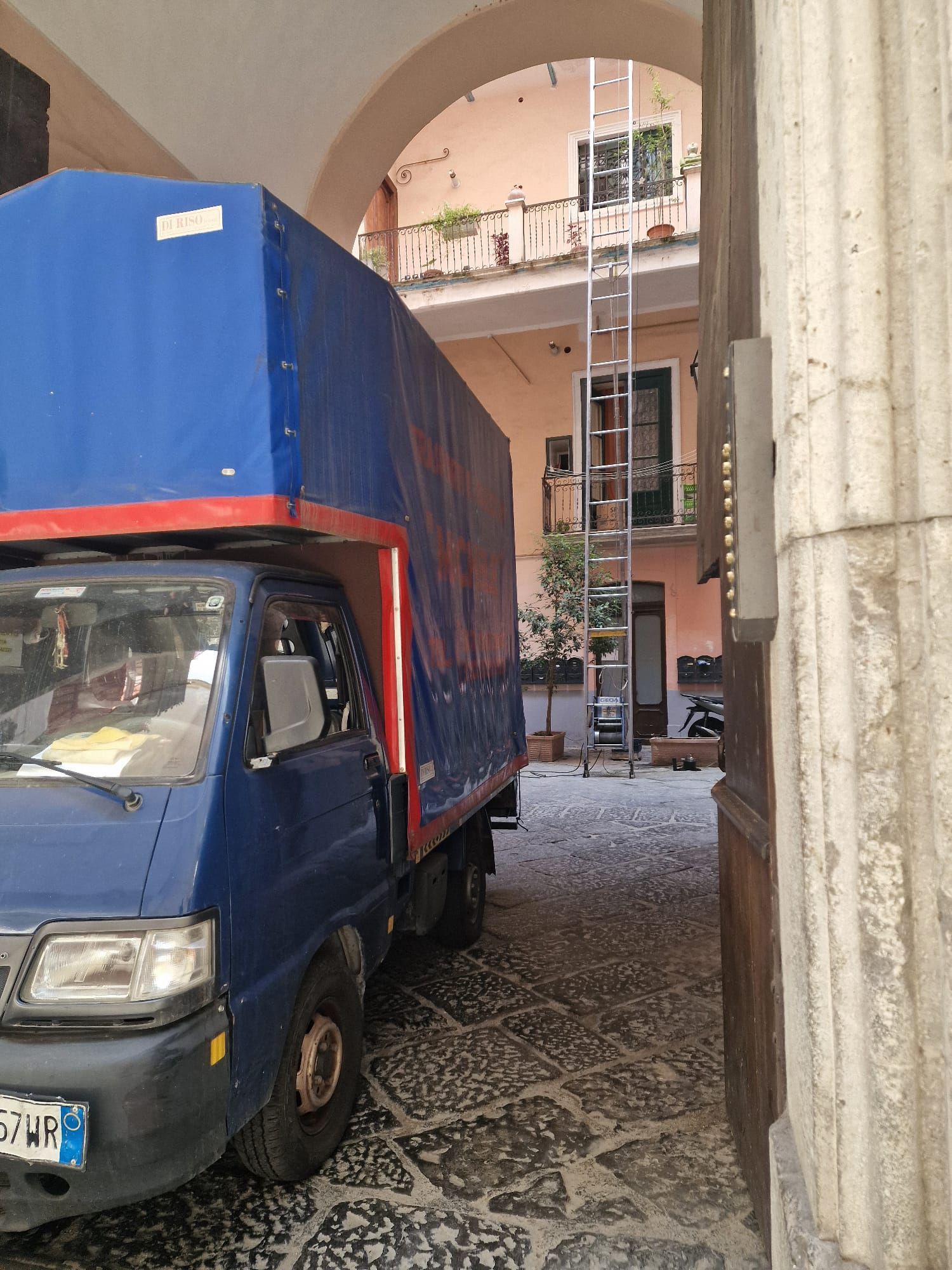 Camion blu sotto un arco di pietra in uno stretto vicolo acciottolato, con edifici e balconi sopra.