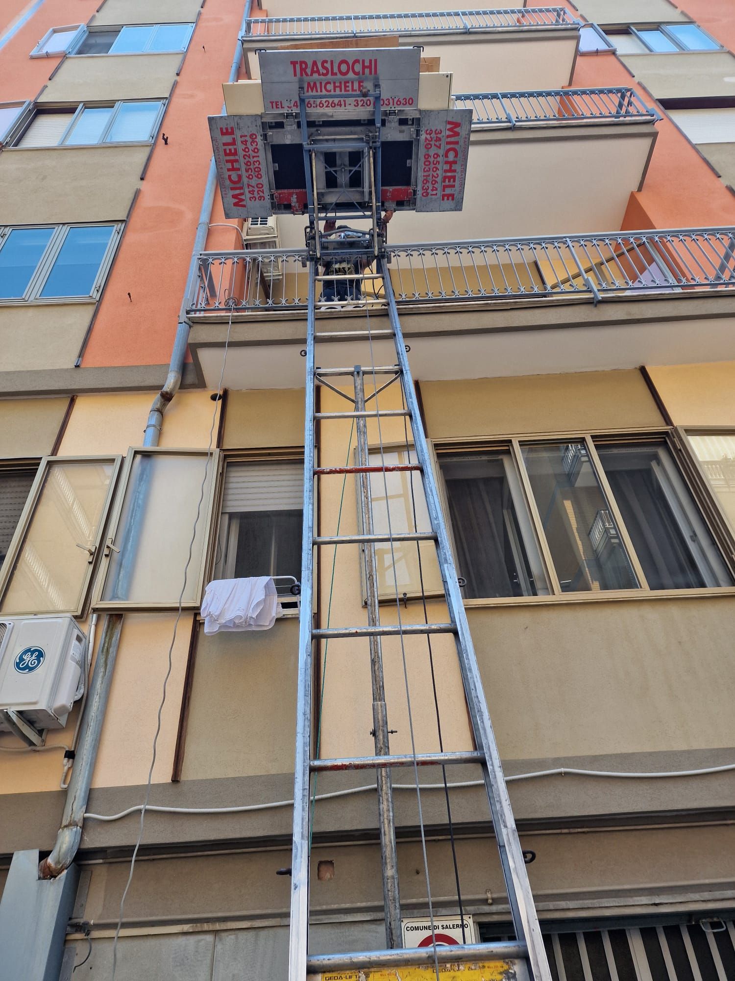 Ascensore di manutenzione adiacente alla facciata del palazzo, che raggiunge la finestra di un balcone con pannelli blu e beige.