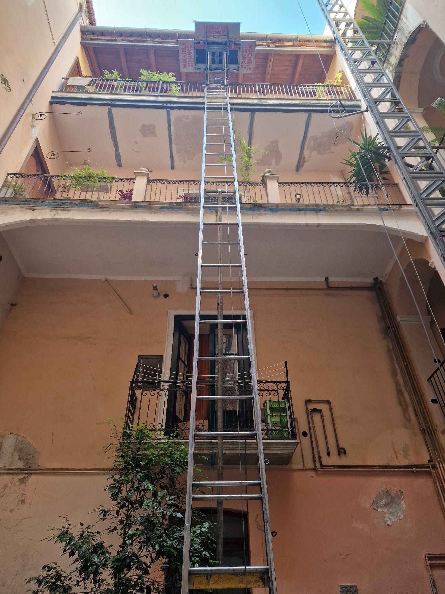 Una lunga scala a pioli si protende verso un balcone al piano superiore di un edificio con cortile interno color pesca.